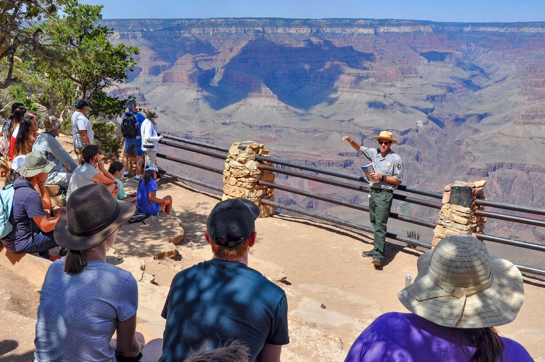 At a scenic overlook with railings, a park ranger is presenting a talk to a group of visitors.