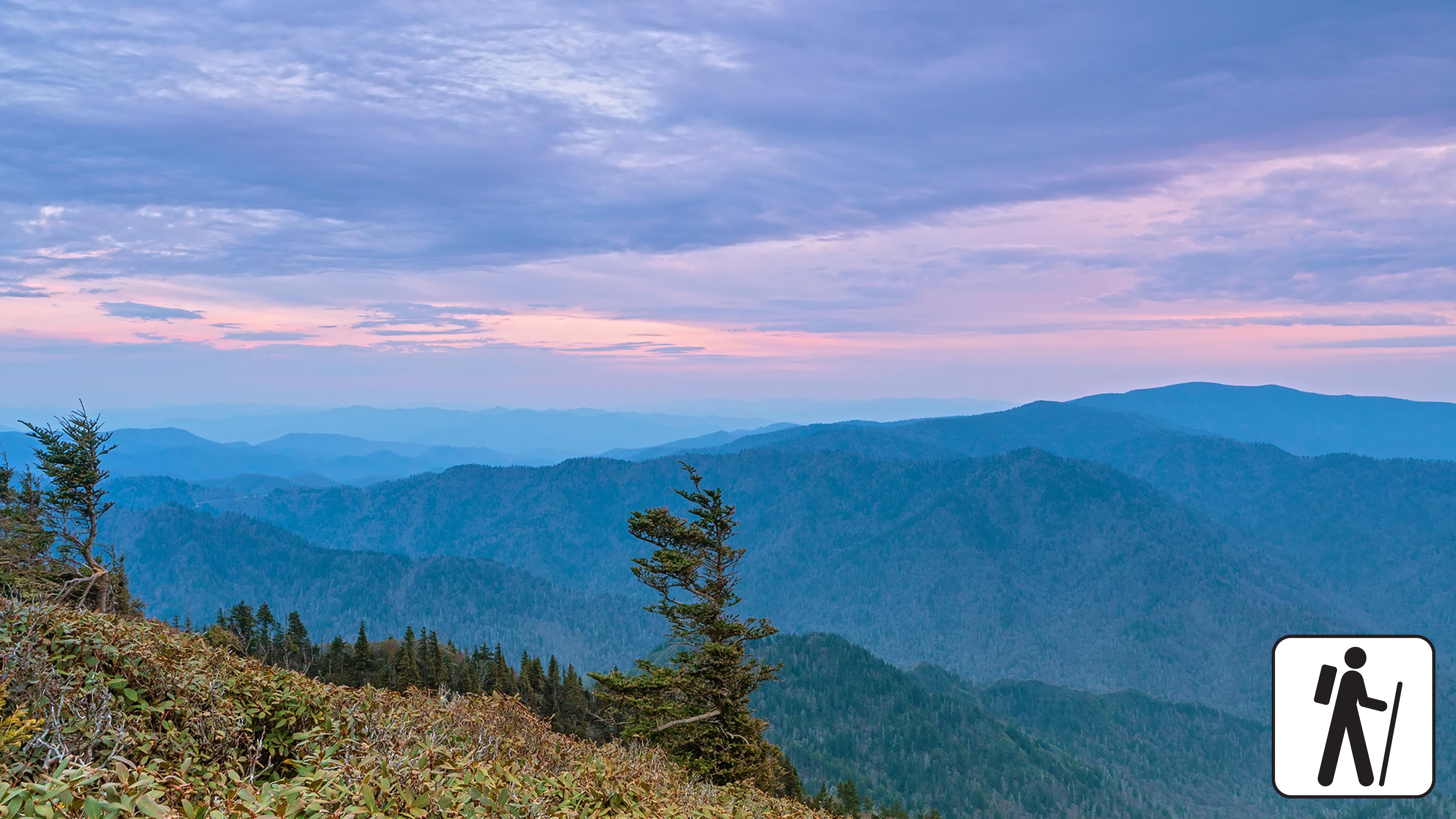 Sweeping mountain views below a pink and purple sky. Hiker icon in corner.