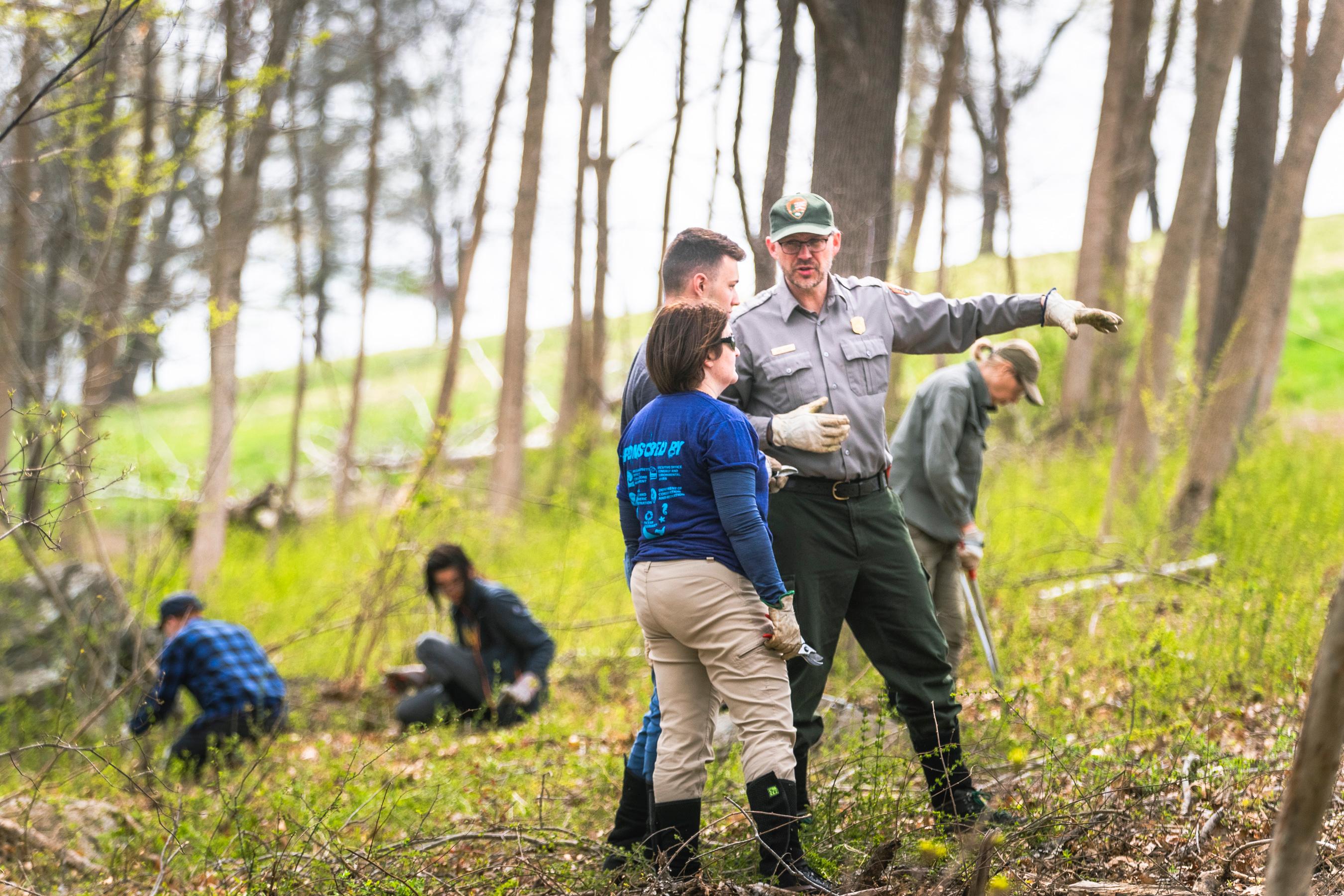 A ranger in uniform talks to a group of volunteers in a grassy and wooded area