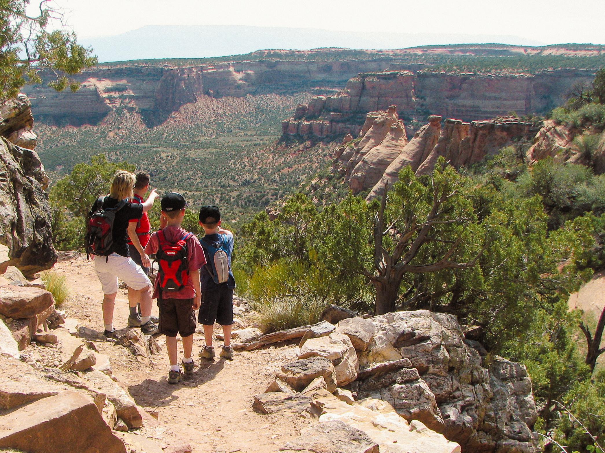 two adults and two children stand on dirt path looking out towards wide open canyons