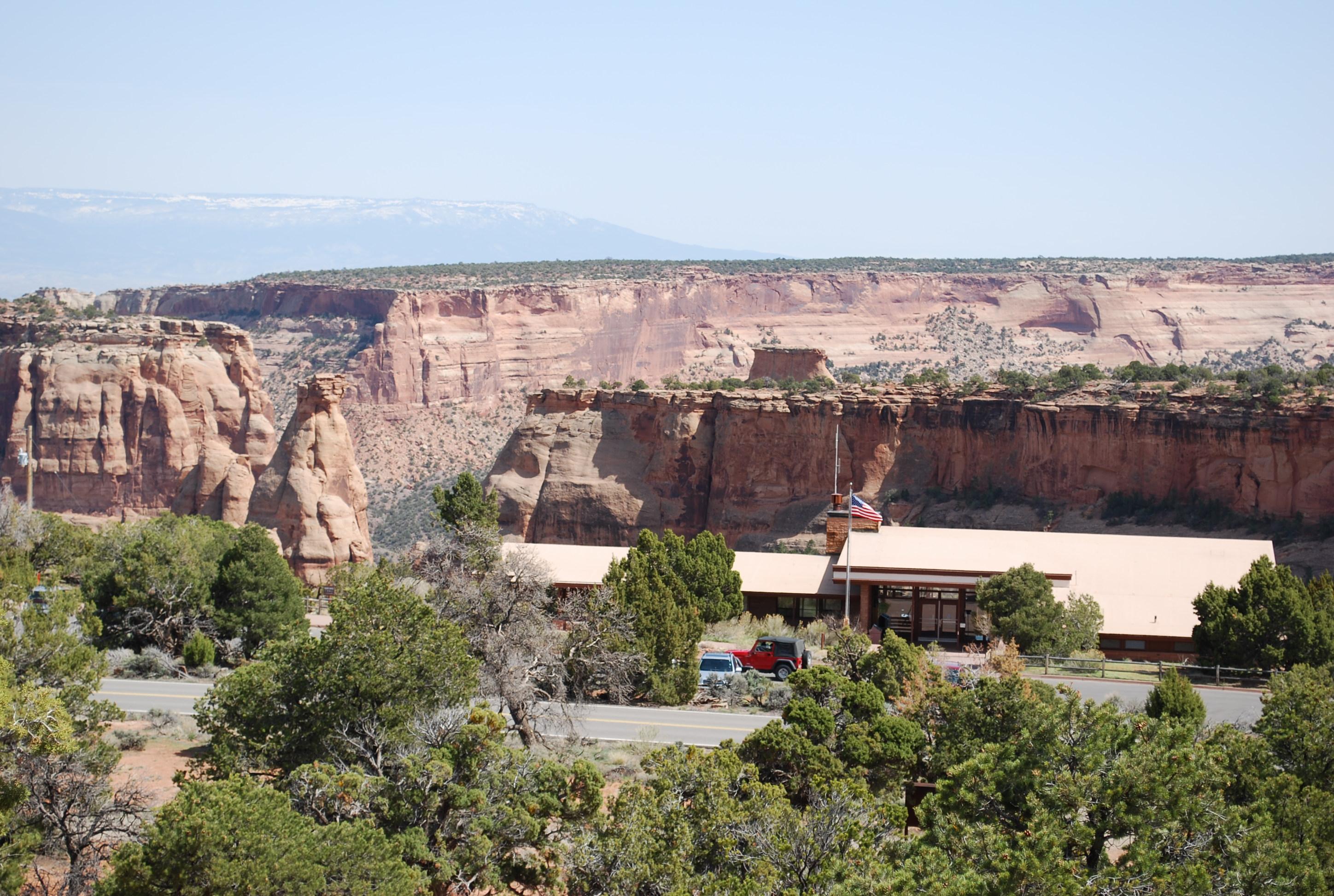 A low, mostly flat building with a flagpole and shrubs, standing before a desert canyon