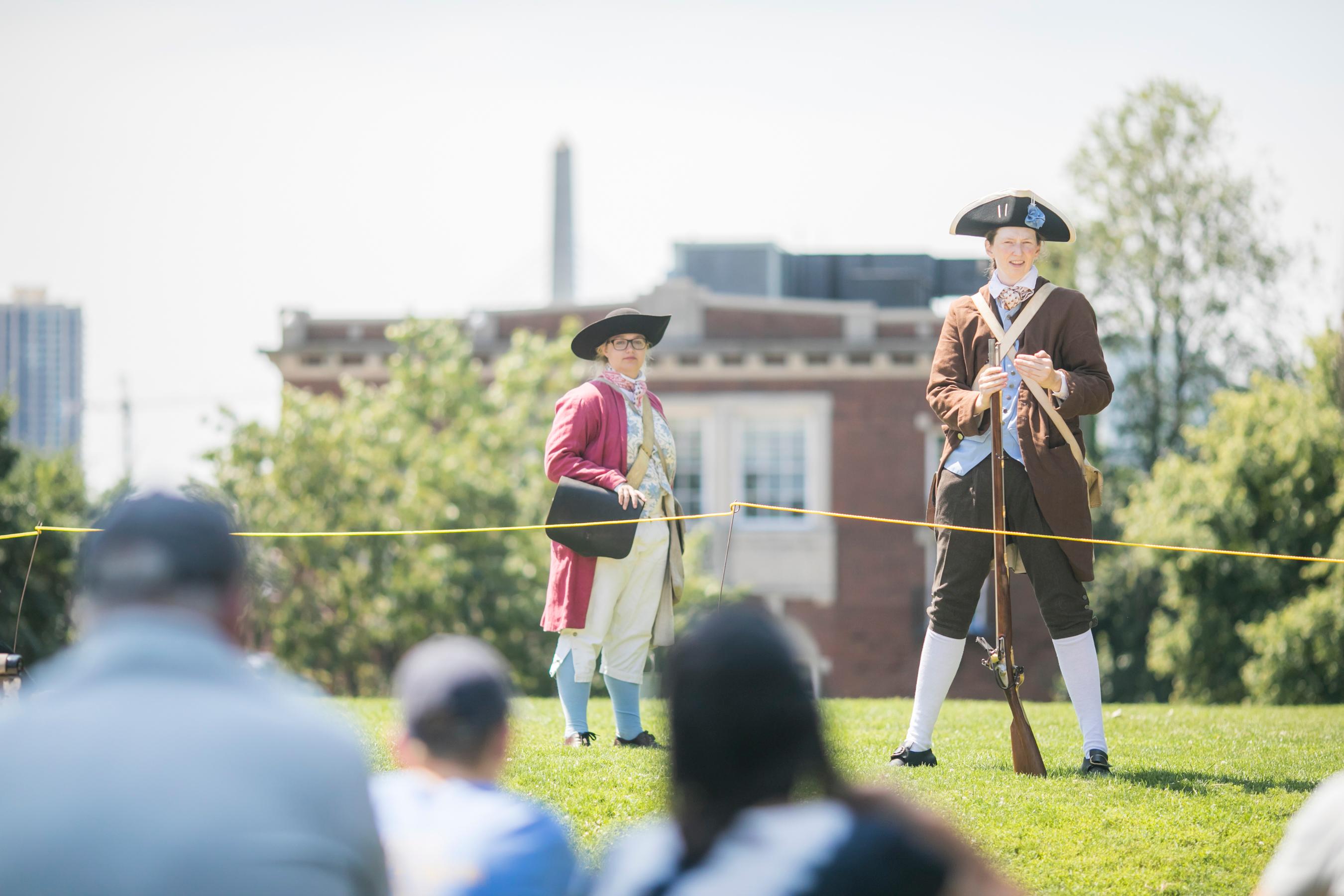 Two Park rangers dressed in period clothing, one has a musket to her side and speaks to the audience